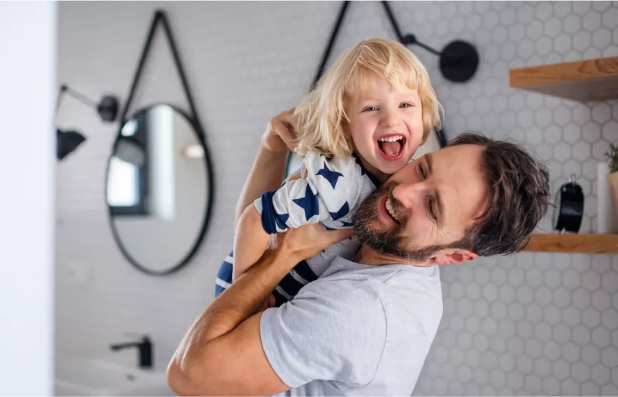 A smiling, bearded man holds a smiling child in his arms in a bathroom